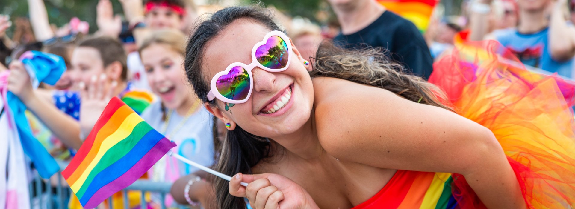 a happy lady dressed in colorful outfit wearing colorful sunglasses holding a LGBTQ+ flag stands out from the crowd during Pride parade.