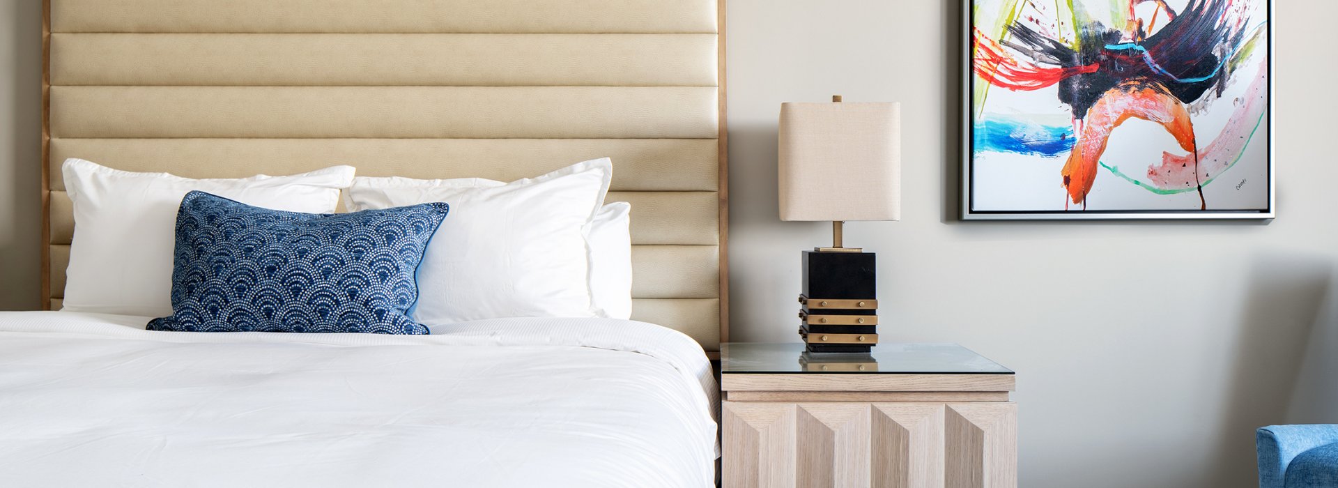 Showing the inside of a hotel room, queen size bed, blue and white carpet, nightstand and abstract painting on the wall.