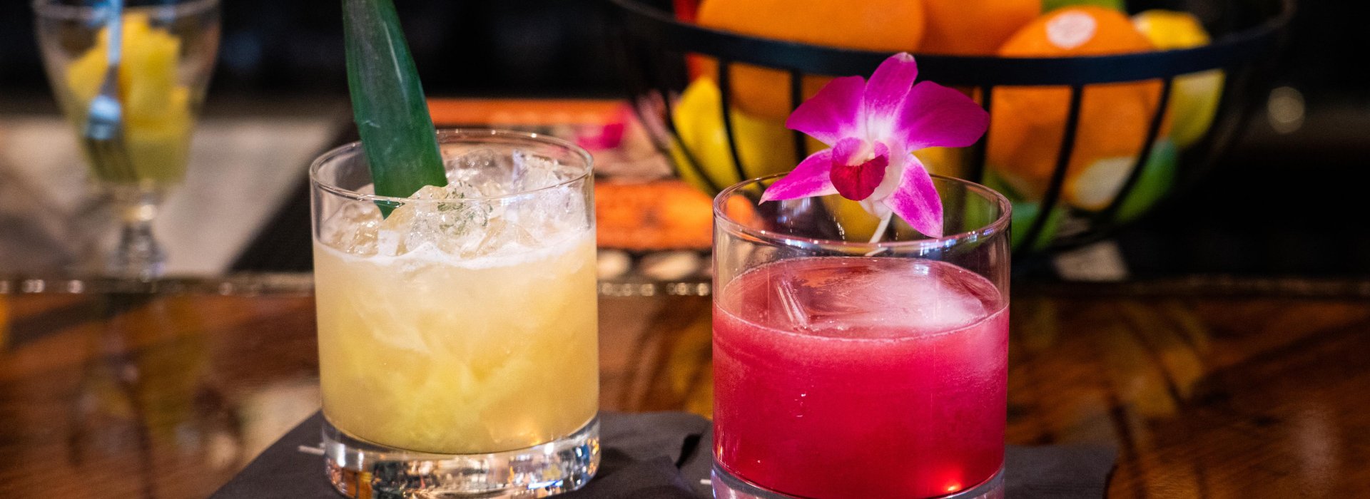 Close up of two cocktail one yellow and one pink in a rocks glass. Basket of fuit in the background.