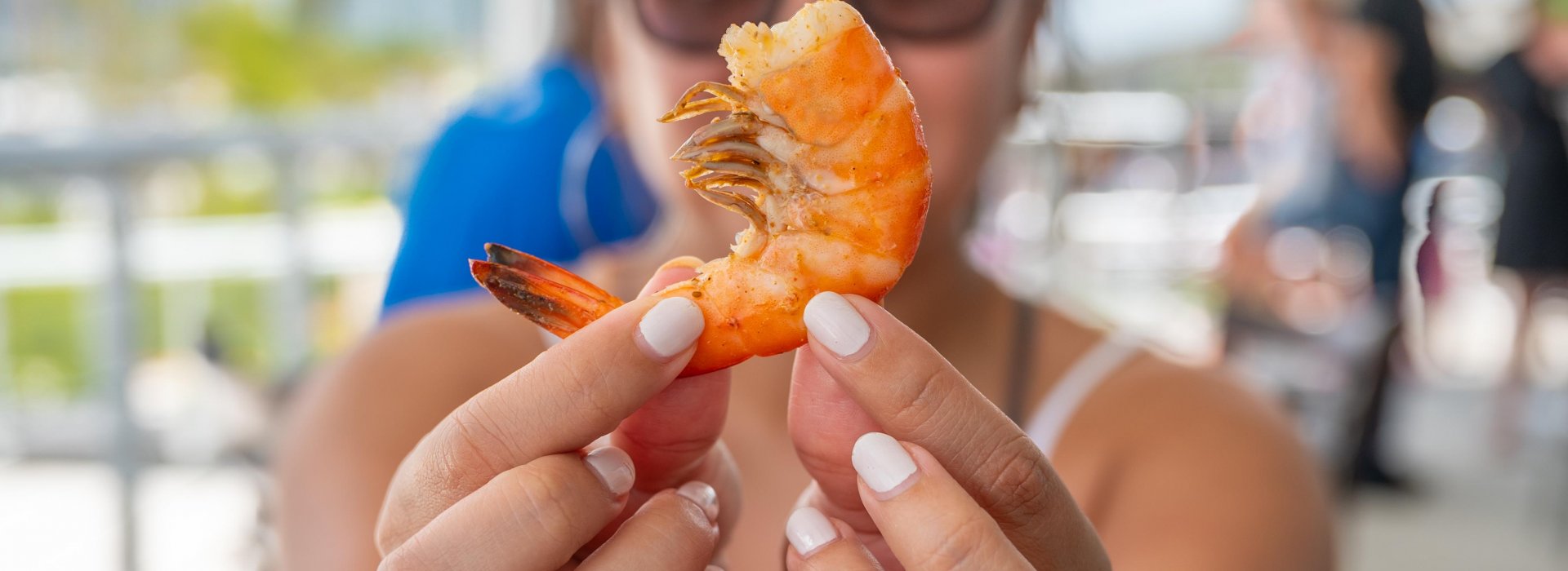 A lady wearing sunglasses holding a shrimp right in front of the camera.