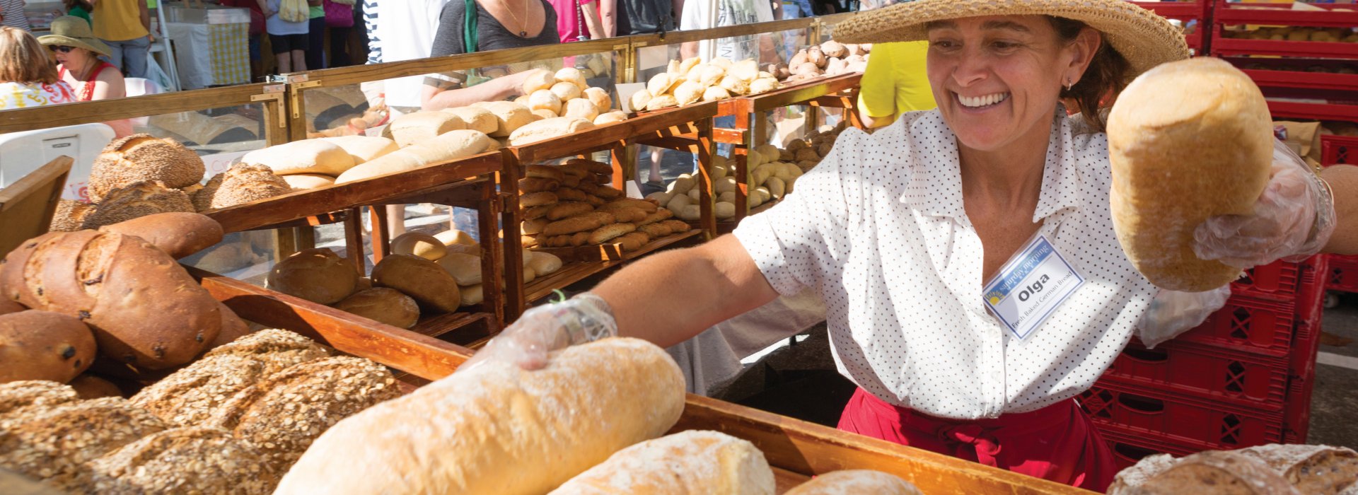 A lady wearing a hat, selling bread with a big smile. Many types of breads are on display.