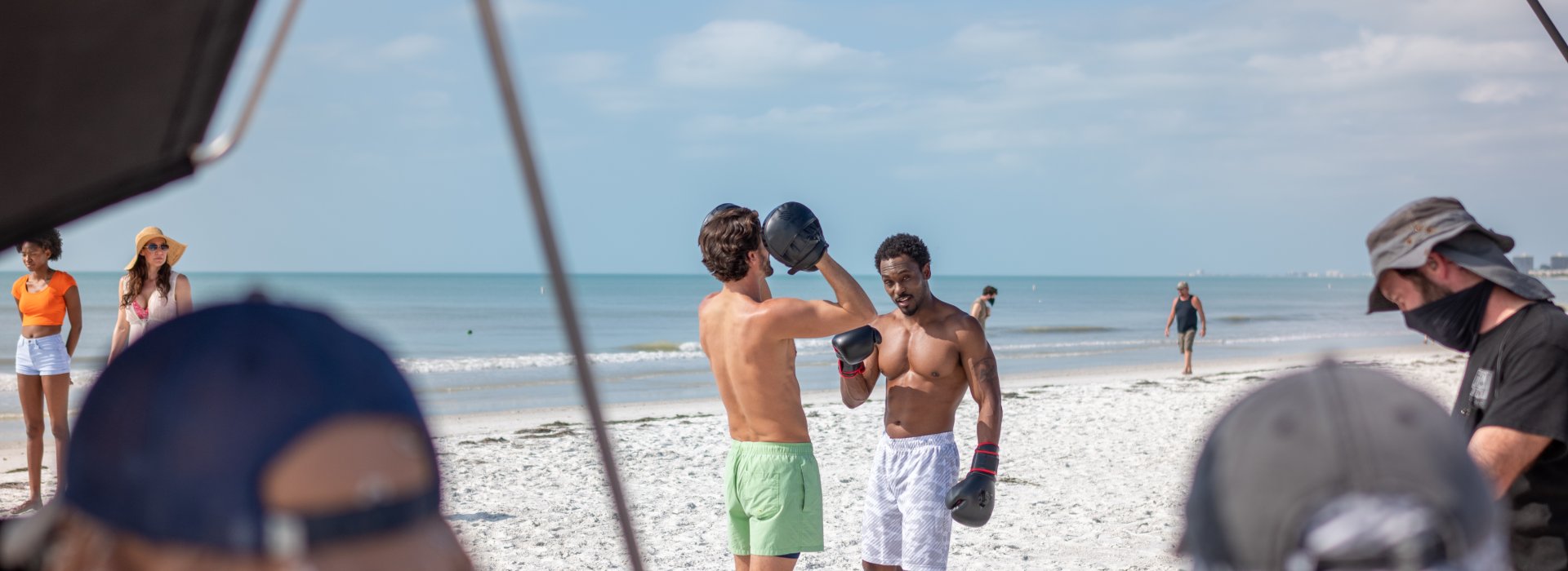 Two men box on the beach in the background with video equipment in the foreground.