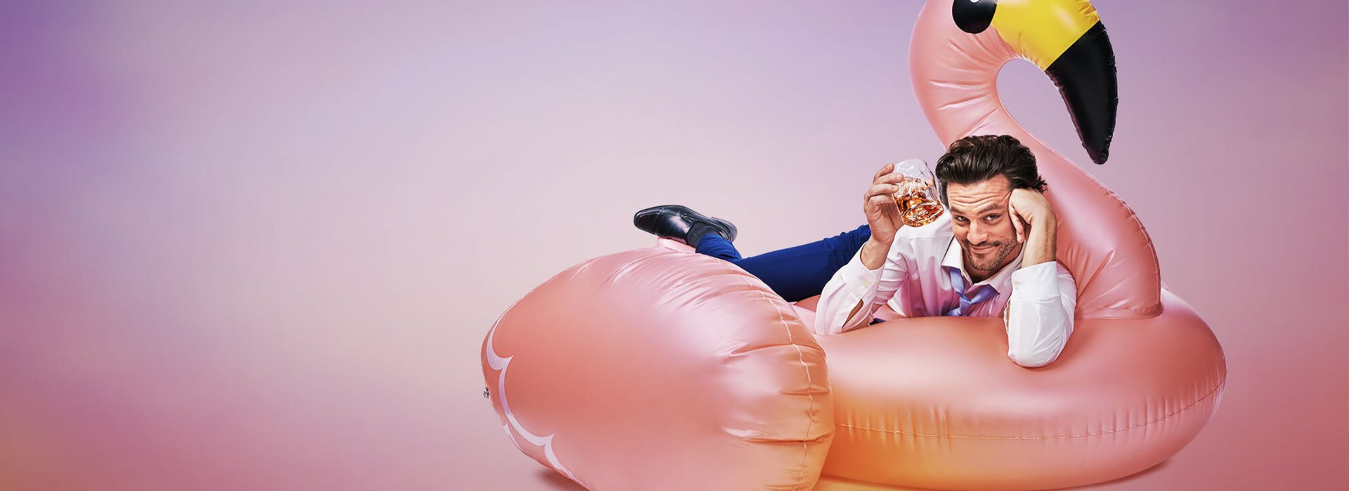 A man laying in a flamingo-floatie holding a glass with liquor agains a pink and purple background.