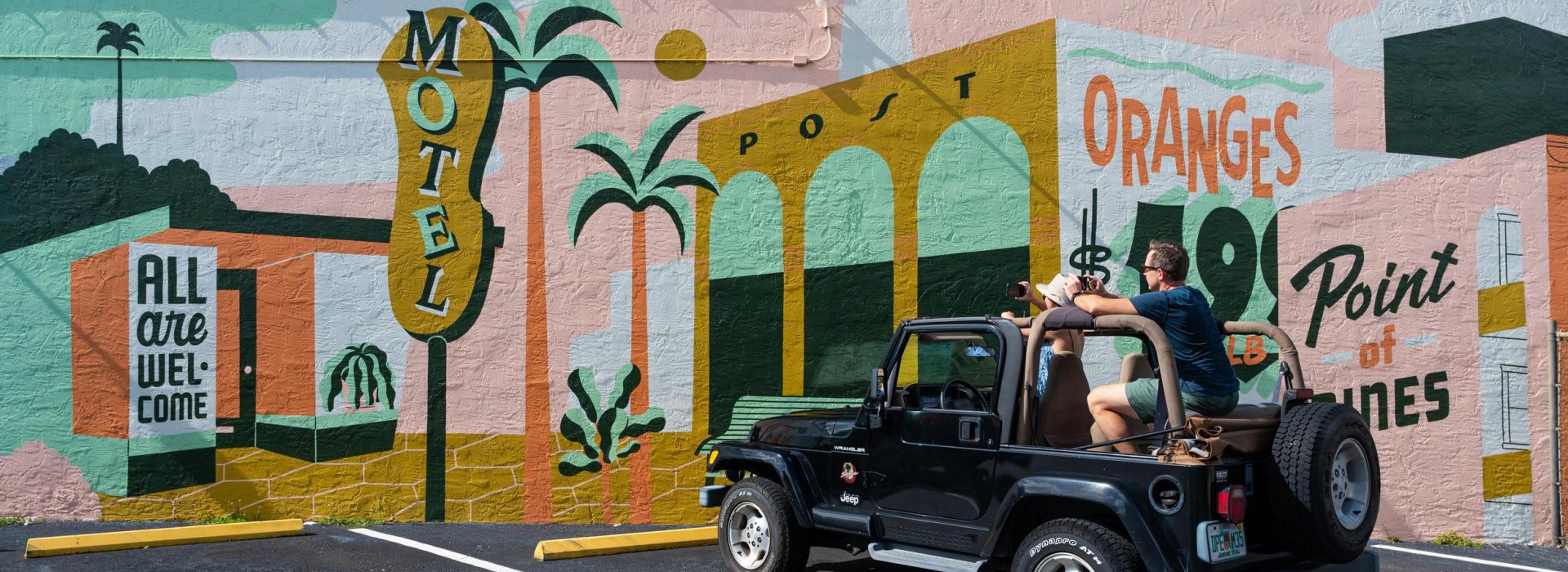 A man and woman in a black jeep park in front of a vintage-themed mural with signs for "motel," "oranges" and "all are welcome here"