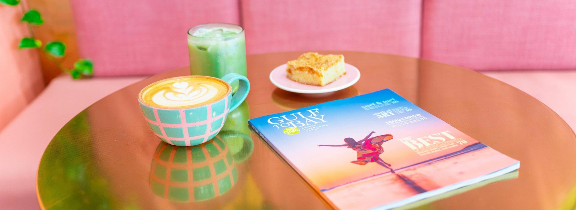 A Destination Magazine sits on a gold colored table next to a latte with latte art, green iced chai tea and coffee cake.