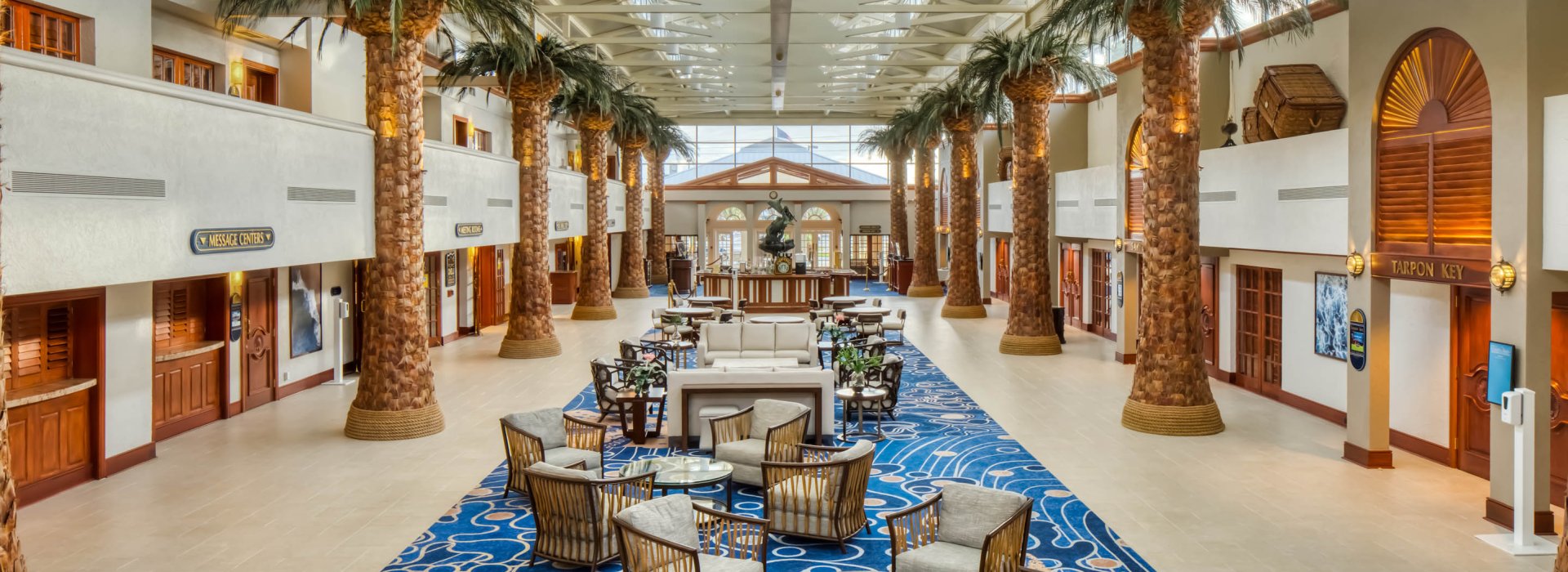 A hotel lobby with chairs and rug down the middle and palm trees flanking the sides