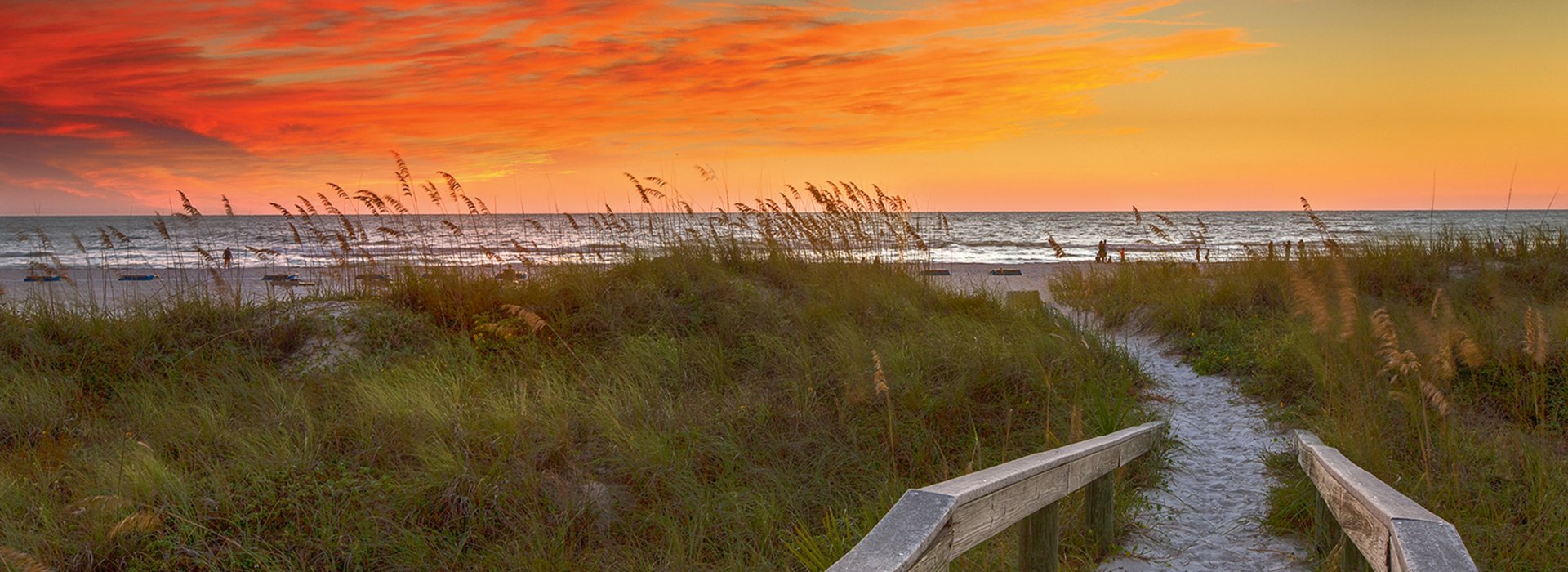 a wooden boardwalk through sea oats and other vegetation leads to the Gulf, the boardwalk is sandy, the sky is just after sunset with orange and blue colors