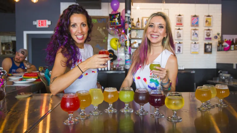 Two servers hold up a beer in front of more beer at Hawthorne Bottle Shop