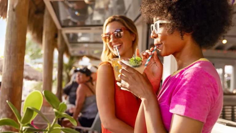 two women in brightly colored clothes sip tropical drinks at a beach bar