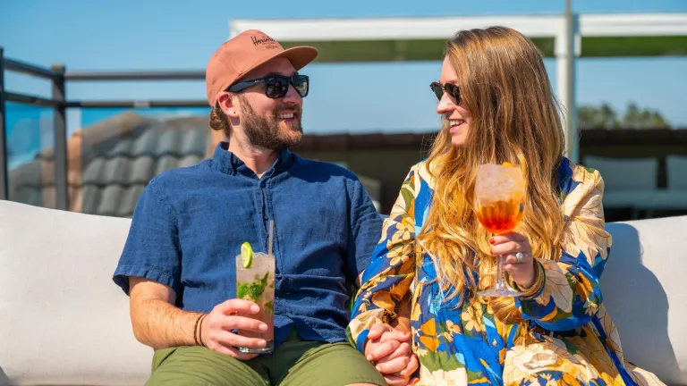 a smiling couple holds hands and tropical drinks on a rooftop at the Hotel Zamora