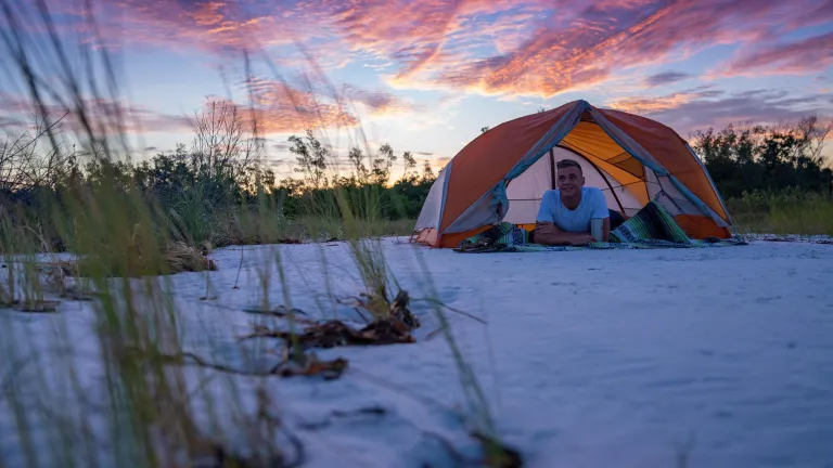 a young man relaxes inside his tent on the beach at Shell key with a dramatic pink and blue sky behind him