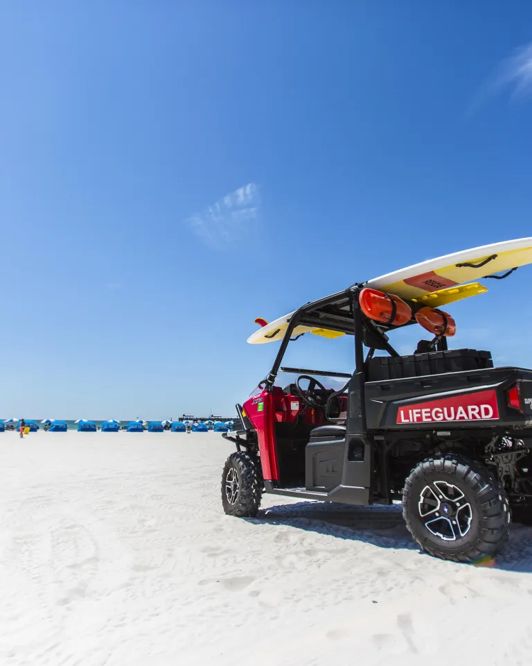 Um buggy salva-vidas estacionado na praia com uma prancha de surfe em cima. Cabanas podem ser vistas à distância.