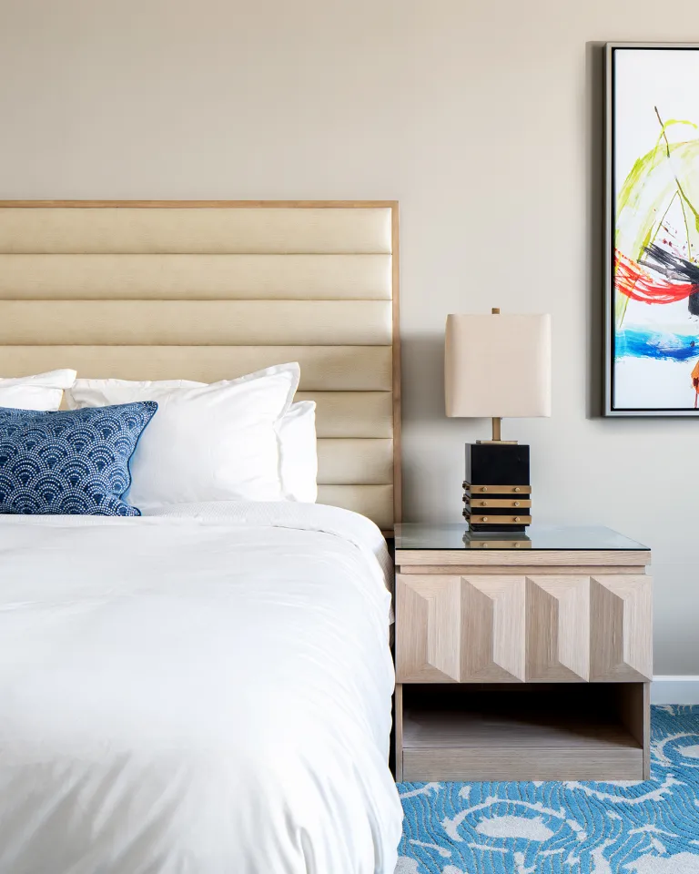 Showing the inside of a hotel room, queen size bed, blue and white carpet, nightstand and abstract painting on the wall. 
