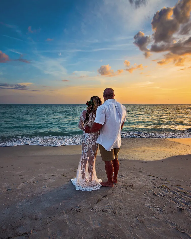 Bride and groom hugging each other and watching the sunset at the beach.