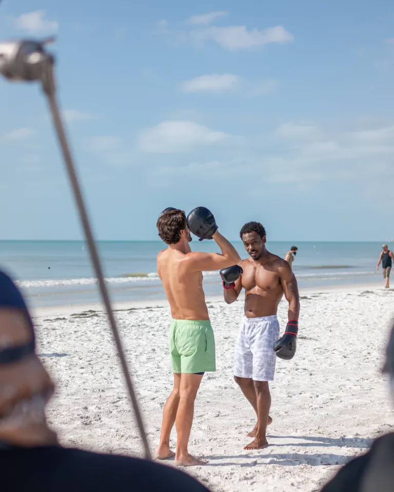 Two men box on the beach in the background with video equipment in the foreground.