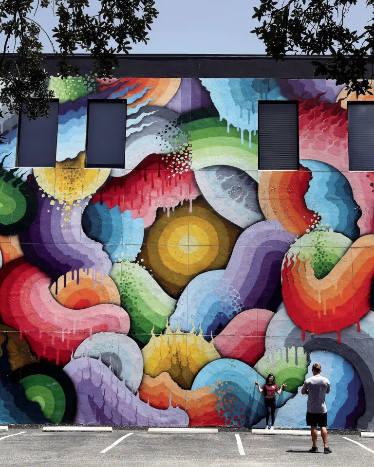 a man takes a photo of a woman in front of an enormous and colorful mural named "Eye of the Storm" in St. Pete's Central Arts District