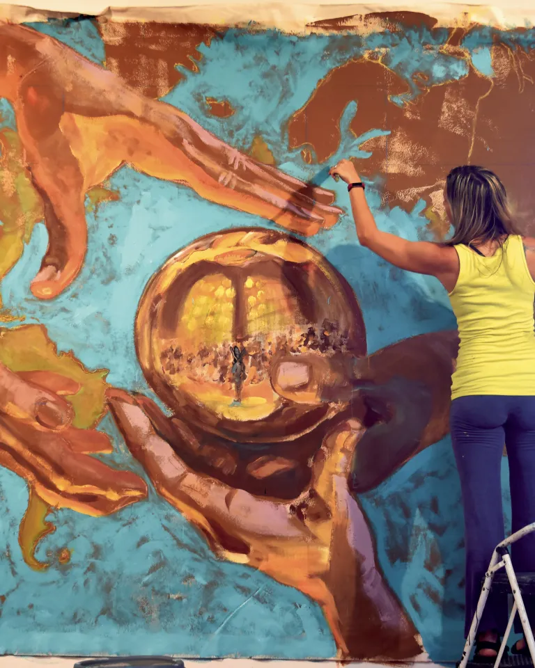 artist Carrie Jadus in a yellow shirt and bluejeans standing on a ladder painting a mural in st. pete
