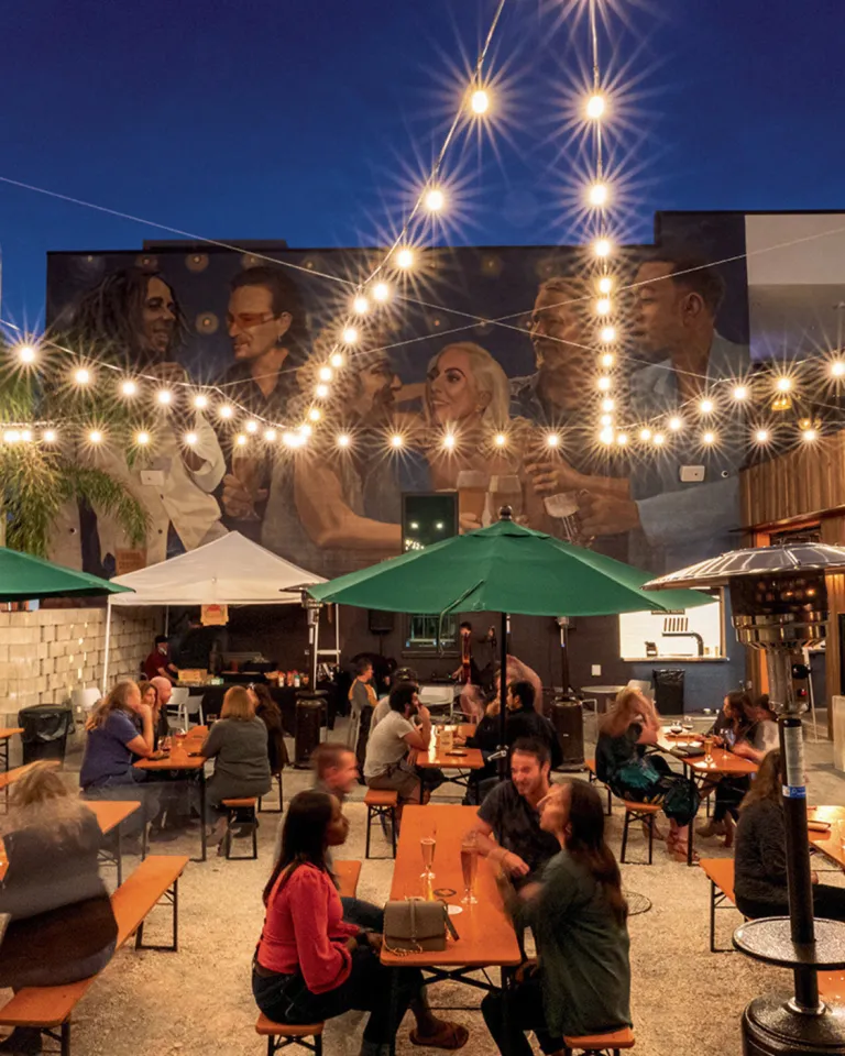 people drinking beer at long picnic tables outside with twinkling lights and a mural in the background at Grand Central Brewhouse