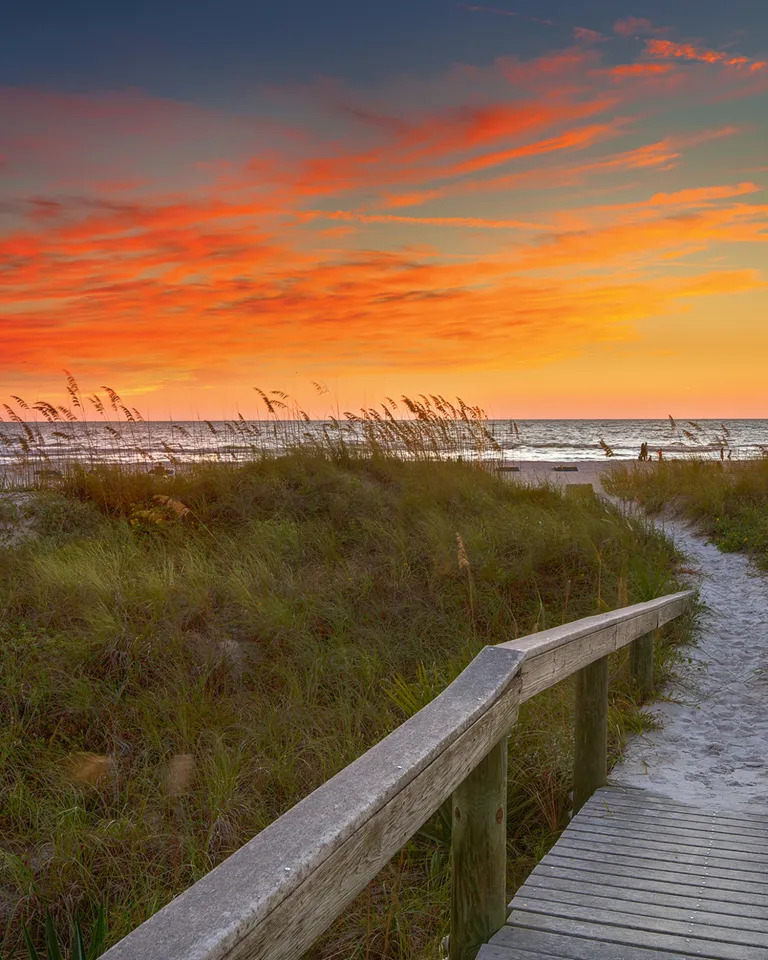 a wooden boardwalk through sea oats and other vegetation leads to the Gulf, the boardwalk is sandy, the sky is just after sunset with orange and blue colors