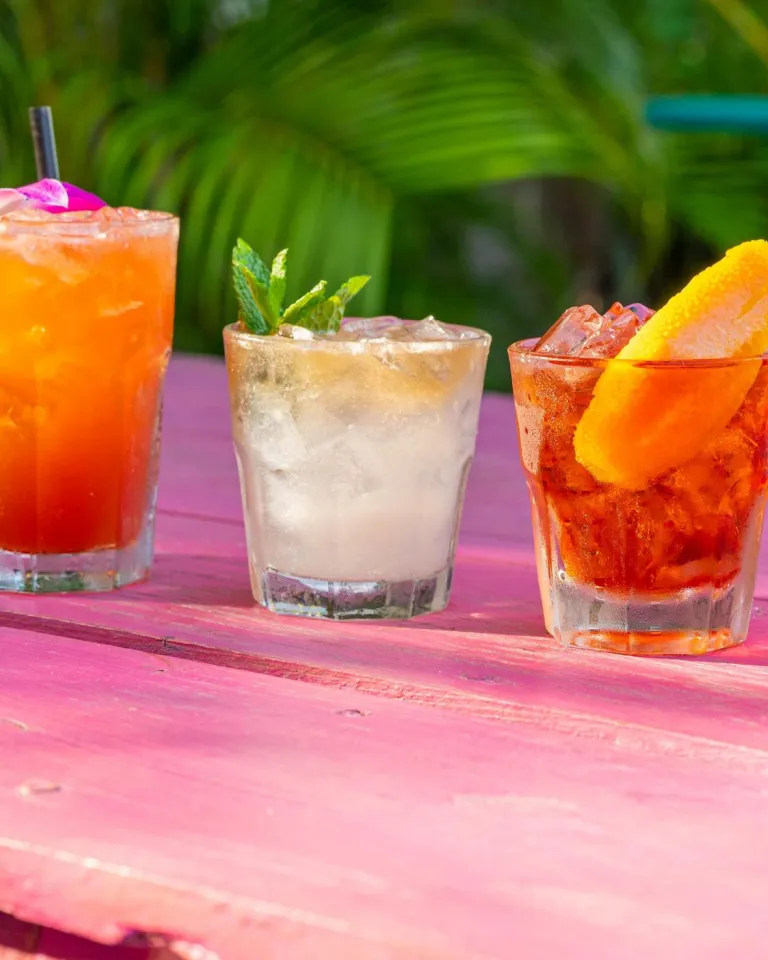 5 cócteles tropicales adornados con frutas sobre una mesa rosa al aire libre