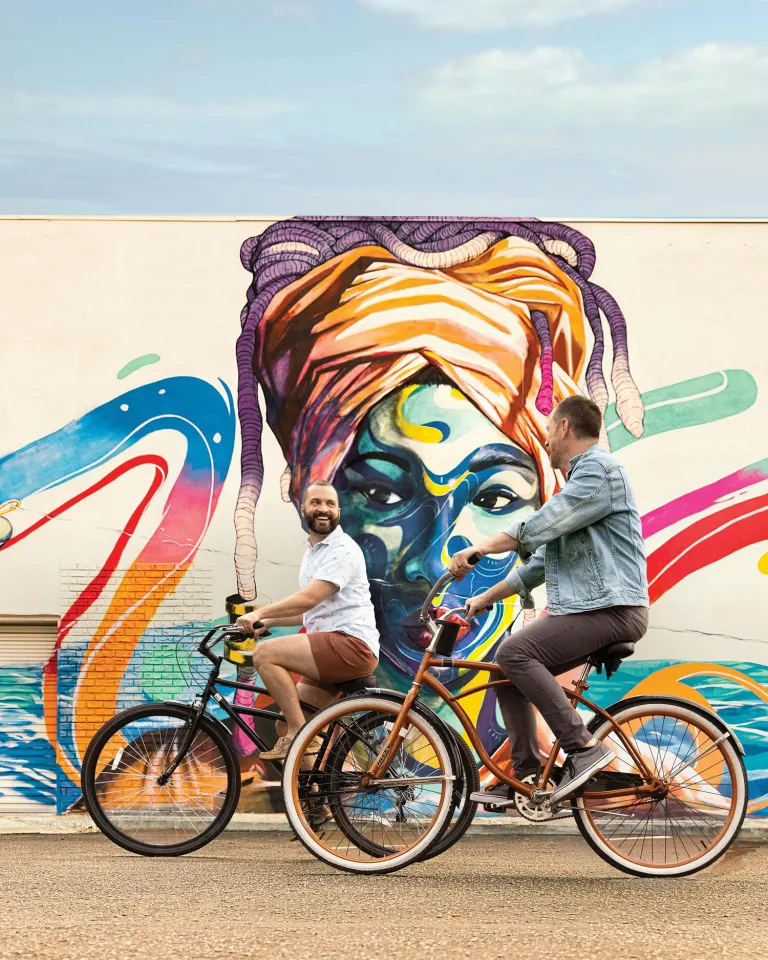 two men riding bikes in front of a colorful mural of an african woman wearing a turban