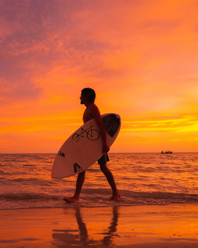 a man with a surfboard walks along the water's edge in Treasure Island at sunset