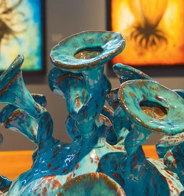 Una obra de arte de cerámica verde azulado y verde en exhibición en el Dunedin Fine Art Center con dos pinturas en la pared detrás