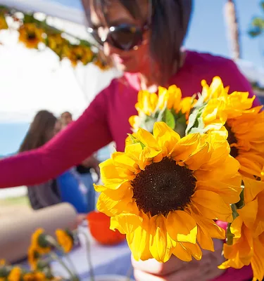 A woman holds a bundle of sunflowers at a sunny, open-air market.