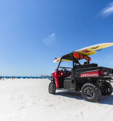 Um buggy salva-vidas estacionado na praia com uma prancha de surfe em cima. Cabanas podem ser vistas à distância.