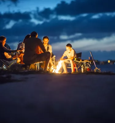 A family circles a bon fire on the beach with a tent in the background