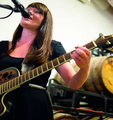 A lady playing a black guitar with beer barrels in the background.