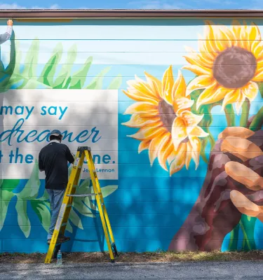 Two people painting hands holding sunflowers and a quote by John Lennon in a mural in St. Pete