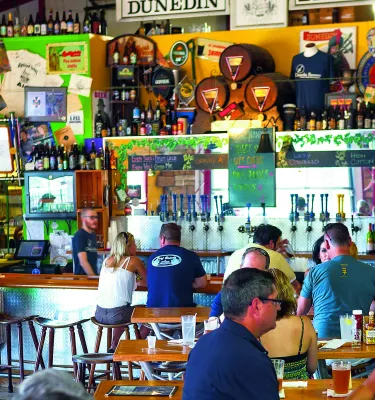 Vista interior de un bar cervecería con gente sentada en la barra y mesas. Pared trasera llena de botellas de osos, parafernalia de cerveza, carteles, instrumentos y más.