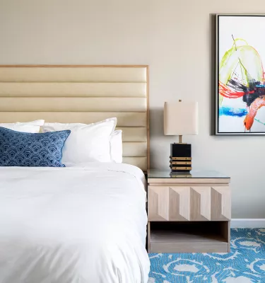 Showing the inside of a hotel room, queen size bed, blue and white carpet, nightstand and abstract painting on the wall.