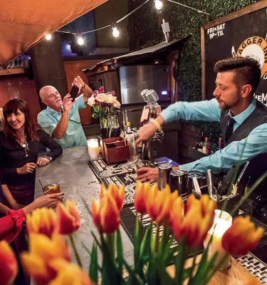 Grupo de personas sentadas en un bar con un camarero sirviendo bebidas. En primer plano hay un jarrón lleno de lirios.