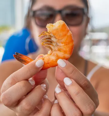 A lady wearing sunglasses holding a shrimp right in front of the camera.