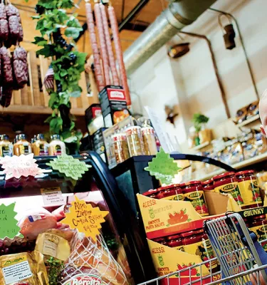 A blonde baby seating in a shopping cart, being fed a cookie. All sorts of cheese, sausage and other cold meats are on display.