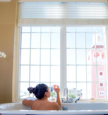 A woman in a bath tub holds a pink champagne glass while looking out the window at the Don Cesar at Spa Oceana