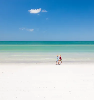 Two beachgoers walk along the white sandy shoreline and emerald green waters of St. Pete Beach on a bright sunny day.