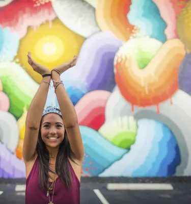 A smiling girl in a red shirt holds her hands up as if to hold the "Eye of the Storm" in a colorful mural by Ricky Watts, Central Arts District