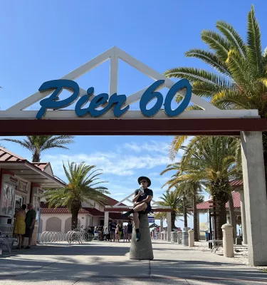 Man sitting under the Pier 60 sign at Clearwater Beach drinking water during the day. Featuring palm trees and sunny skies.