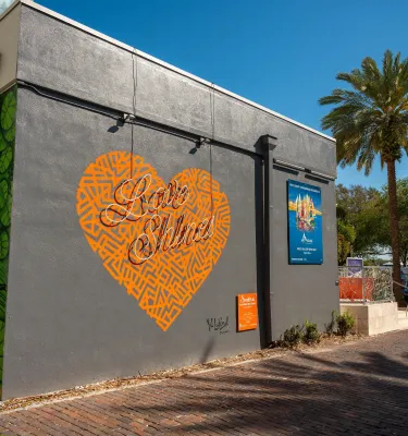a mural of an orange heart that says Love Shines on a dark gray building with a blue sign and a palm tree to the right