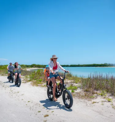 Uma mulher e um bebê em um passeio de bicicleta elétrica ao lado de água azul-esverdeada e aveia do mar com outros membros da família andando de bicicleta atrás deles no Fort De Soto Park