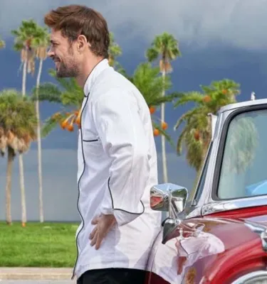 A man and a woman stand facing each other outside next to a red car with palm trees in the background.