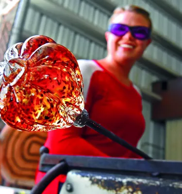 A woman holds an orange piece of art glass in a hot shop