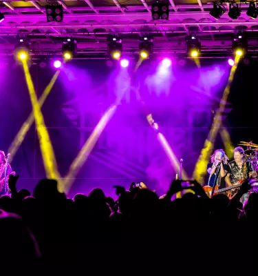 Outdoor concert at night featuring yellow and purple lights, a crowd of people and a band playing on stage.