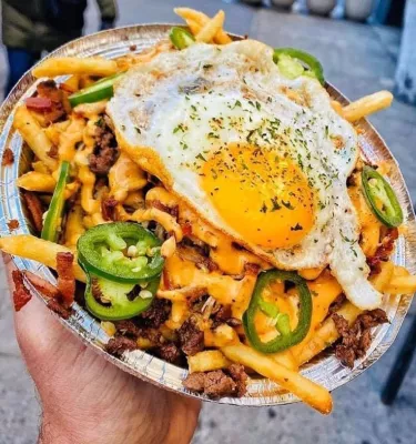 Photo of a hand holding a bowl of cheesy french fries with egg and Jalapeño peppers on top.