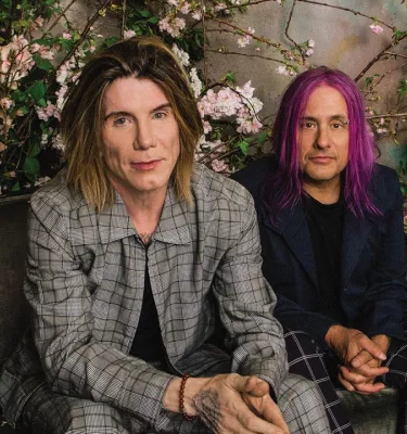Musicians Goo Goo Dolls staring at the camera will sitting in front of trees and plants.