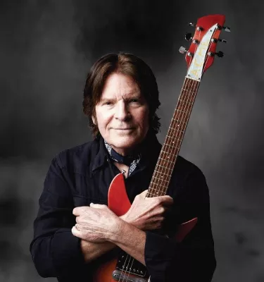 Musician John Fogerty holding a red electric guitar and standing in front of a dark and cloudy sky background.