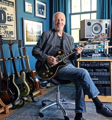 Musical artist Peter Frampton sitting in a chair with a guitar in a music room surrounded by various guitars.